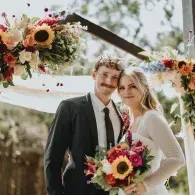 Two people in wedding attire holding a colorful bouquet under a floral arch outdoors