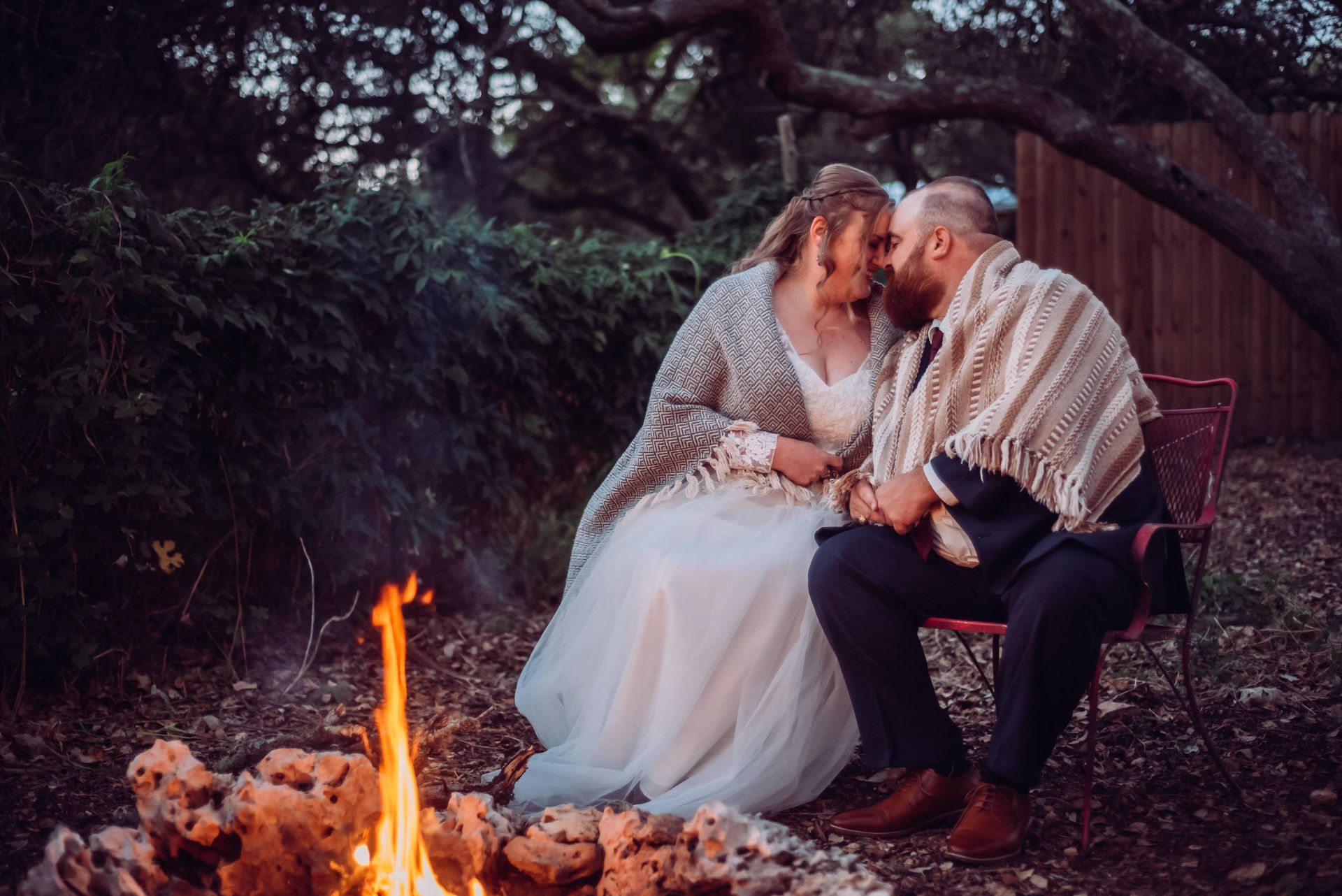 Couple sitting by a campfire in a wooded setting, leaning close and smiling at each other.