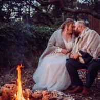 Couple in wedding attire leaning close by a campfire in a wooded setting at dusk