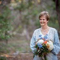 Woman in a light blue dress holding a colorful bouquet outdoors, smiling in a wooded setting