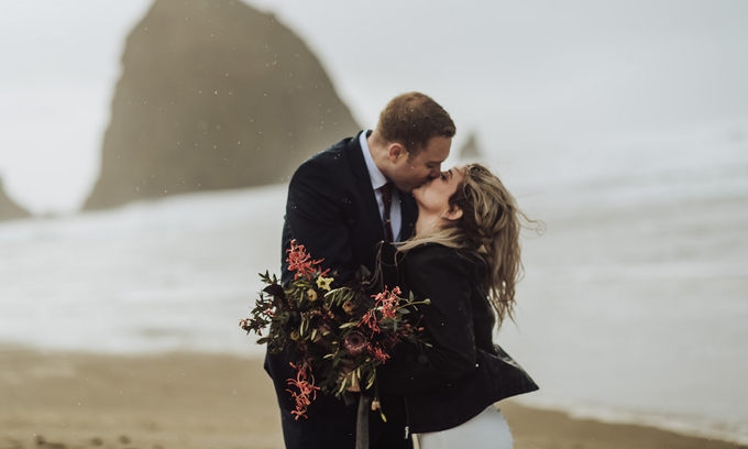 Couple embracing on a foggy beach, holding a bouquet near a large rock formation