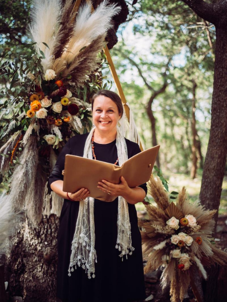 Woman holding an open book beside a decorated tree outdoors in a wooded setting