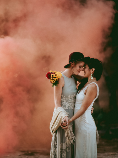 Two brides in white dresses and black hats kissing beside a bouquet amid pink smoke