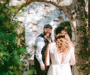 Bride faces smiling man beneath a stone archway in a garden, with greenery framing the scene