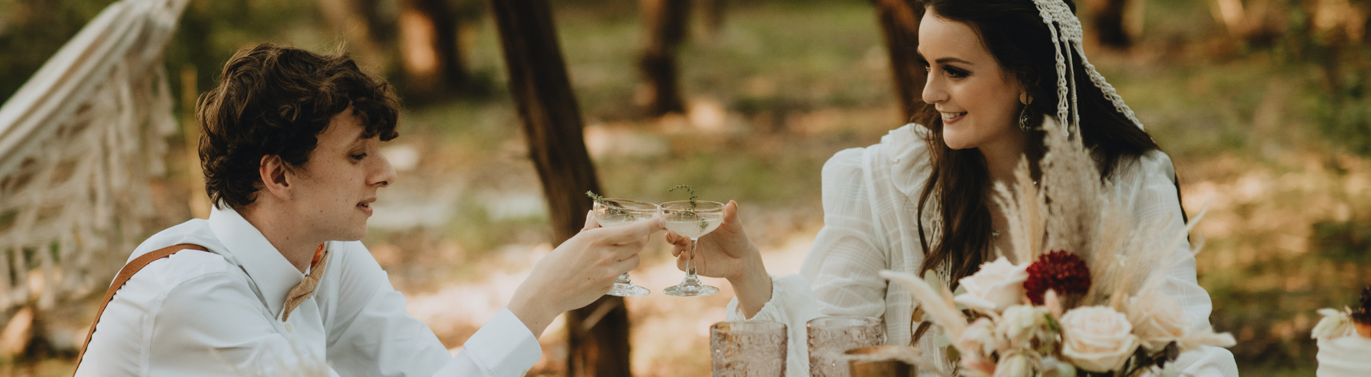 Two people in white outfits toasting drinks at an outdoor table with flowers in a warm, wooded setting