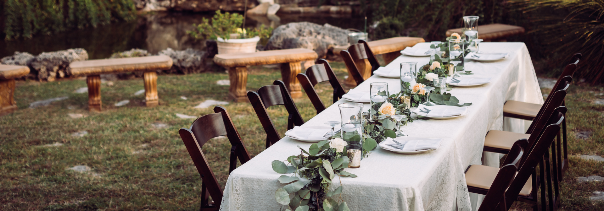 Long outdoor dining table with white tablecloths and greenery set beside a rustic garden wall