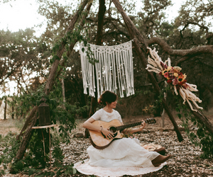 Person in white dress playing guitar outdoors under hanging macramé decorations.