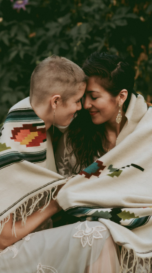Two people wrapped in a cream blanket, touching foreheads outdoors in soft light.