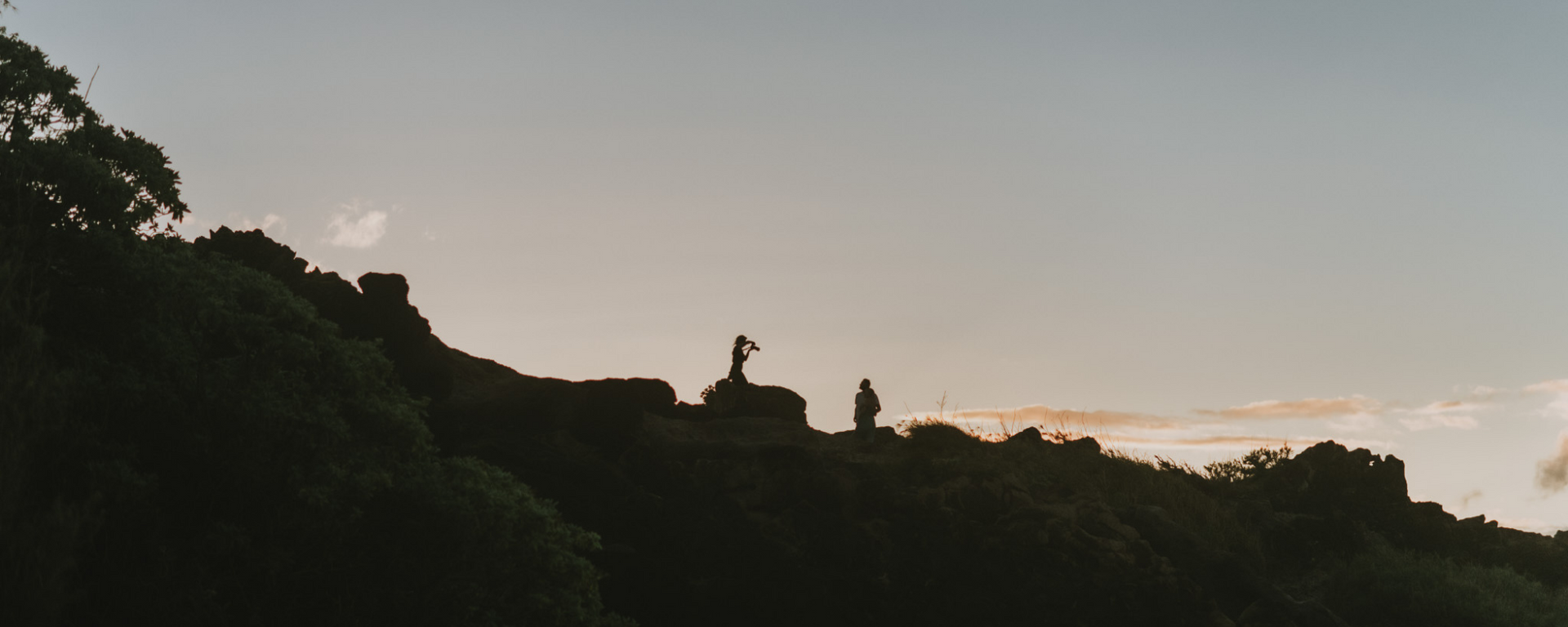 Silhouetted figures on a hill at sunset under a cloudy sky, with dark trees framing the scene