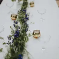 White table with a floral runner and gold-rimmed glasses at a place setting, with a wine glass nearby