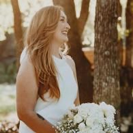 Smiling woman in a white dress holding a bouquet outdoors by trees