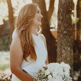 Smiling bride in a white dress holding a white bouquet outdoors among trees