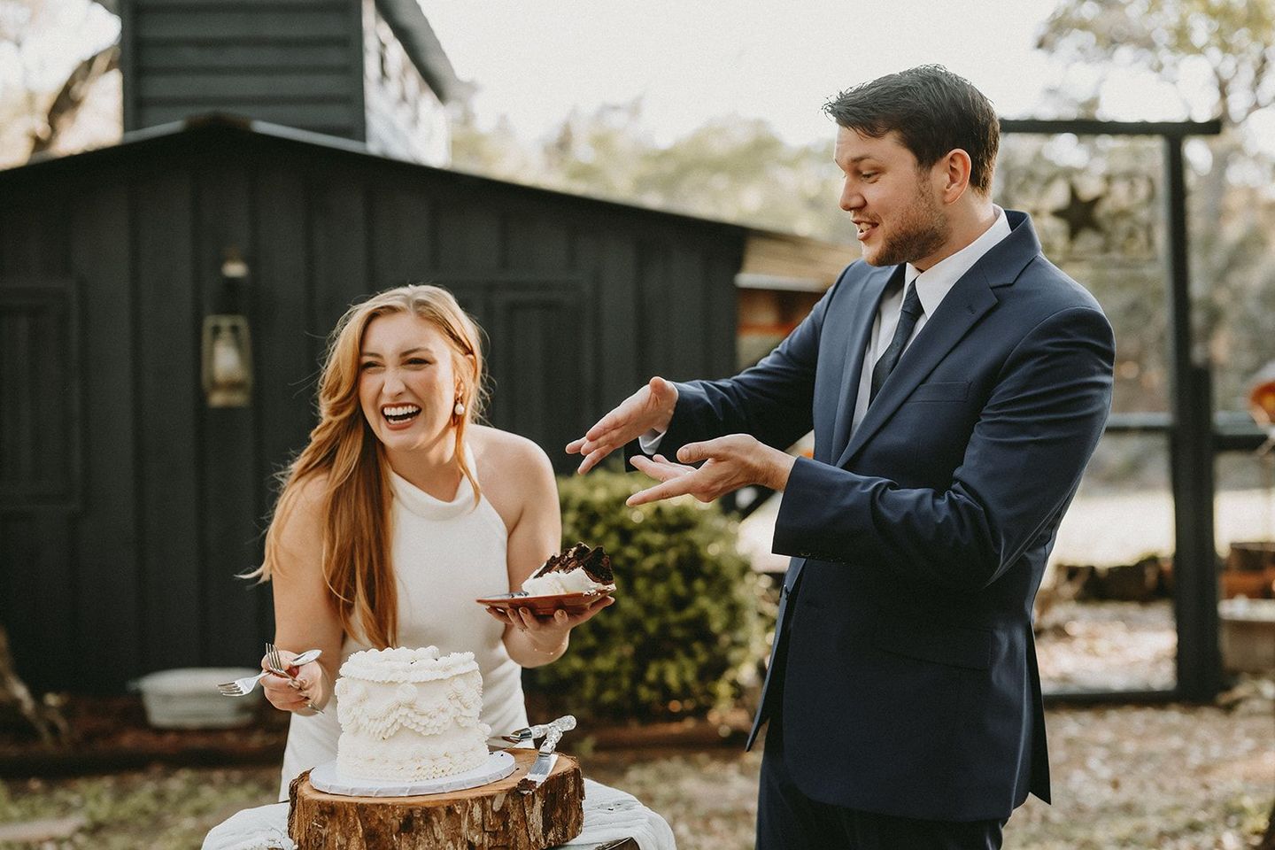 Bride and groom cutting a wedding cake outdoors, both smiling by a rustic black cabin