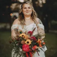 Woman in white dress holding a colorful bouquet outdoors, with soft greenery and blurred lights behind her