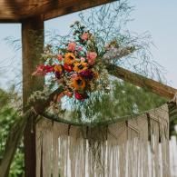 Colorful floral arrangement hanging beneath a wooden beam with fringe decor and greenery backdrop