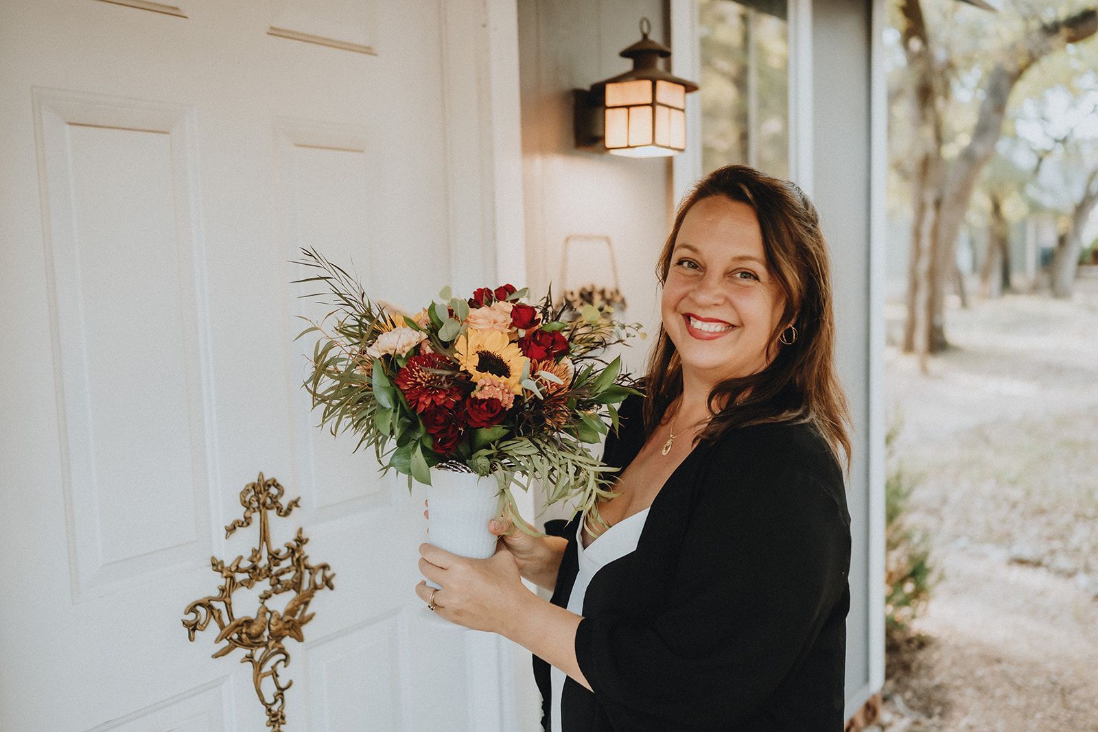 Smiling woman holds a bouquet by a white front door with a decorative brass knocker.