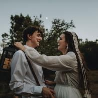 Two people embracing outdoors at dusk, smiling at each other near trees and a lit window.