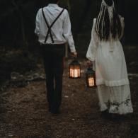 Two people in white walking through a dark wooded path, carrying lanterns