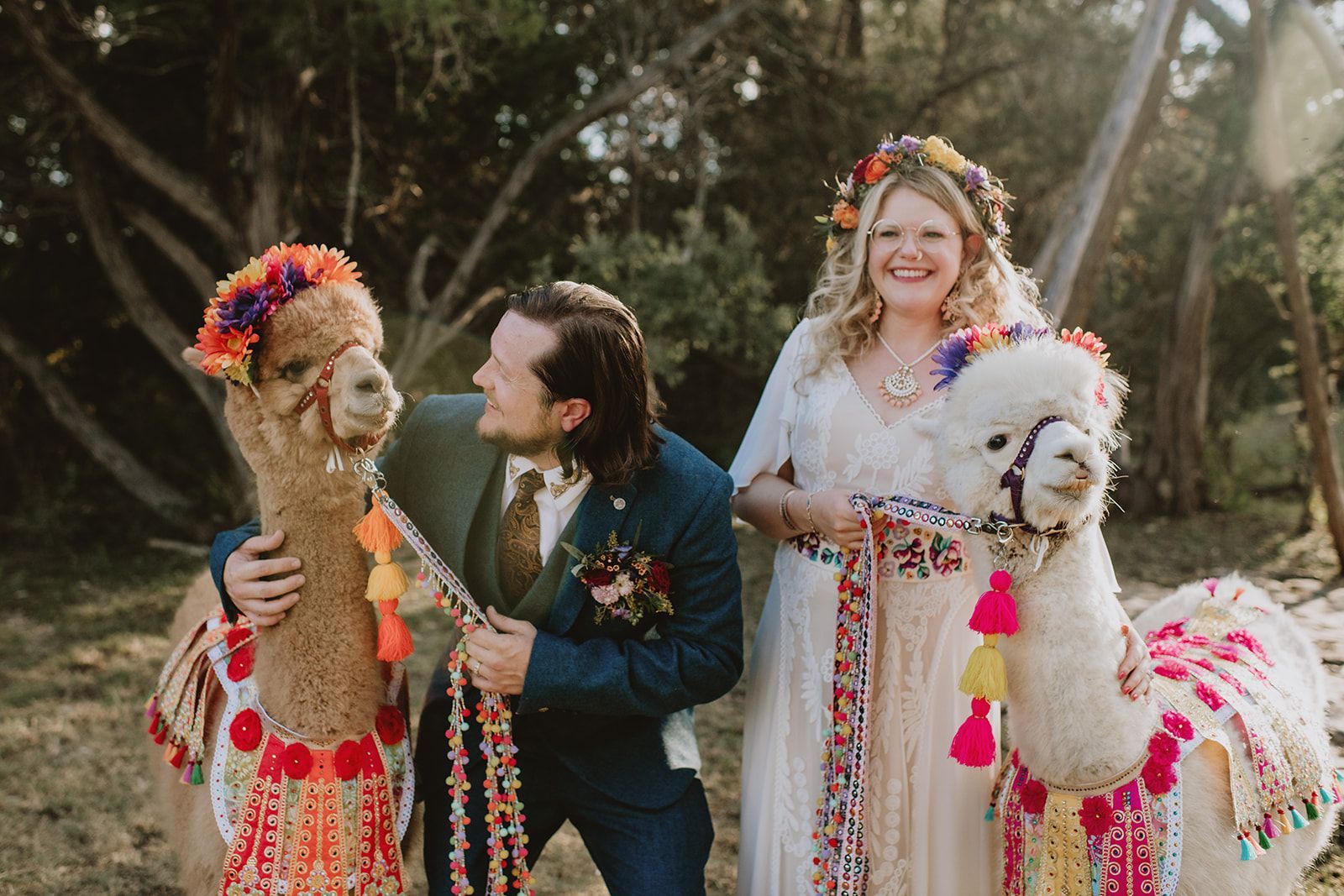 Bride, groom, and two decorated llamas in a sunlit outdoor wedding scene, smiling and standing together