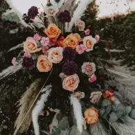 Floral arrangement with peach, pink, and dark purple roses, white feathers, and greenery