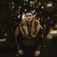 Person in a fur-trimmed coat standing outdoors among trees in soft, dark light