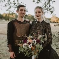 Two people in dark formal outfits holding a bouquet outdoors, smiling beneath a tree.