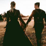 Two people in dark clothing holding hands and walking through a field at sunset