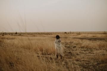 A person in a white coat stands in a dry grassy field under a cloudy sky.