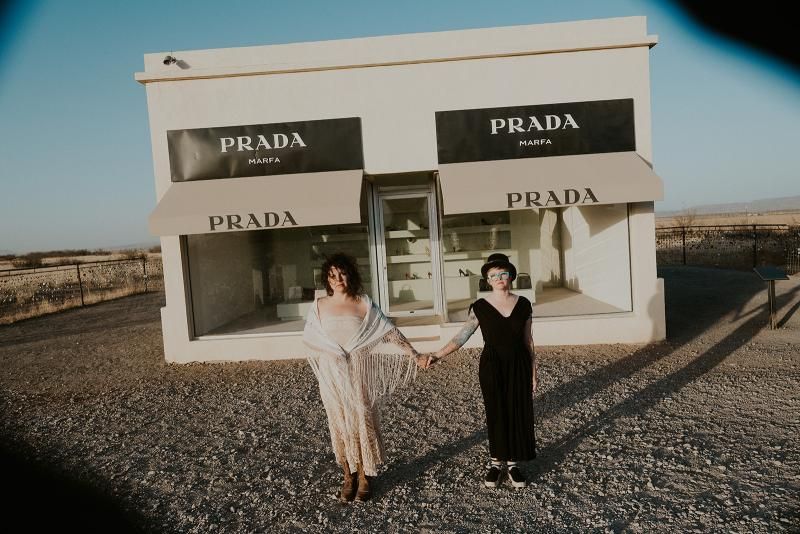 Two people posing outside a small white Prada storefront in a desert-like setting.