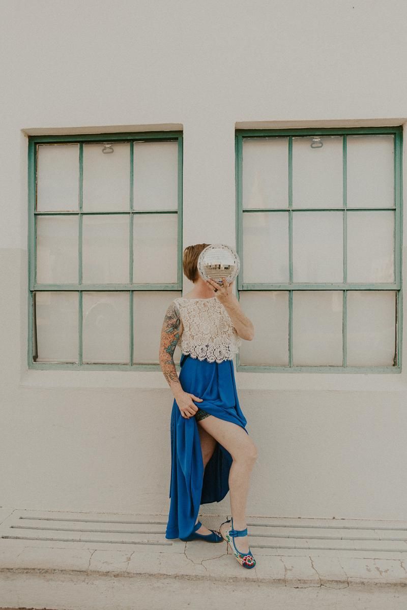 Person in a blue skirt and lace top poses against a wall between two windows.