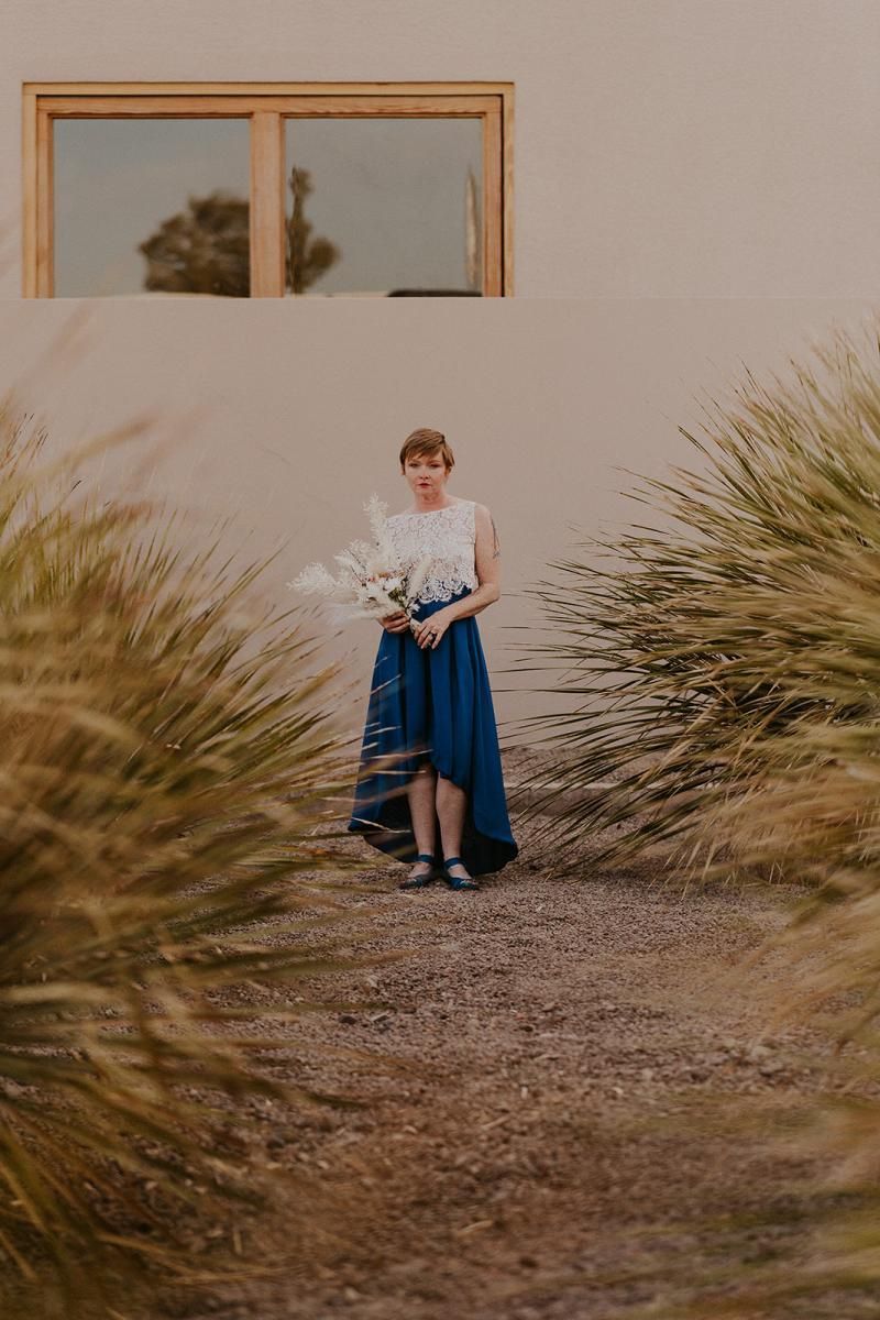 Person standing on a gravel path between desert grasses, holding flowers in front of a beige building.