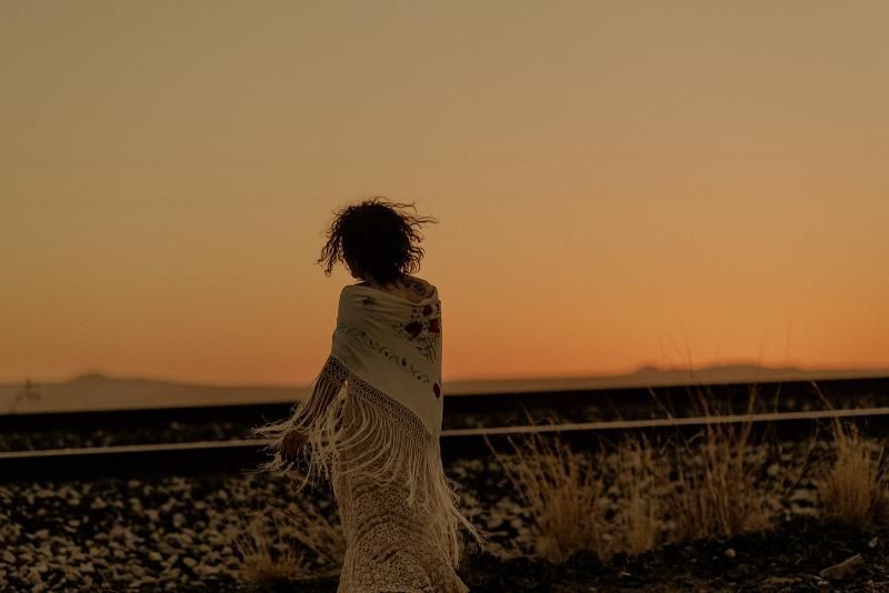 Person wrapped in a blanket stands by railroad tracks at sunset, silhouetted against an orange sky.