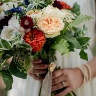 Hands holding a bouquet of cream, peach, white, and red flowers with green leaves