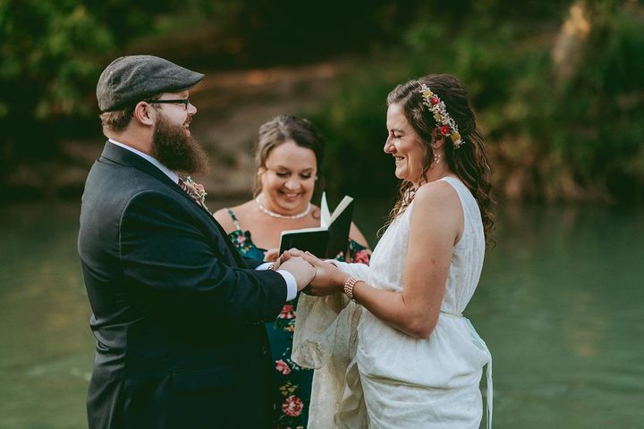 Couple exchanging rings during an outdoor wedding ceremony by a river, officiant smiling behind them