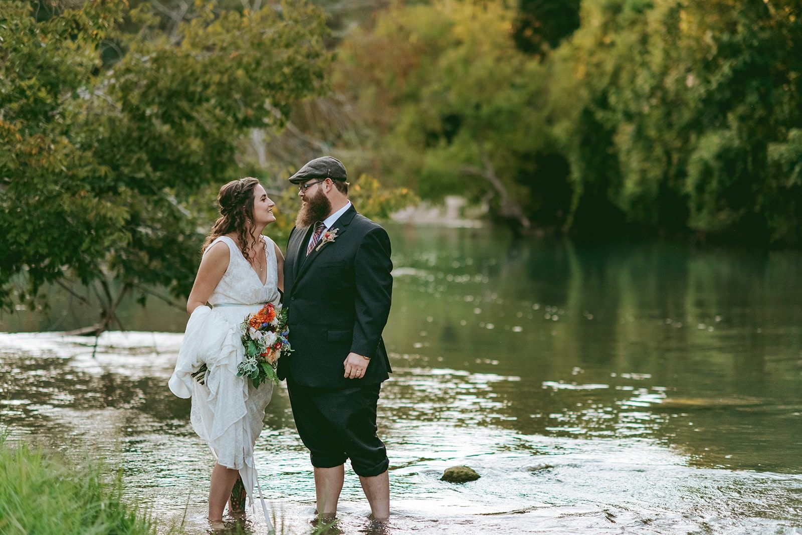 Couple in wedding attire standing barefoot by a river, smiling in a leafy outdoor setting