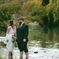 Couple in wedding attire standing in shallow water by a lake, surrounded by greenery.