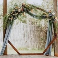 Floral wedding arch with draped gray fabric and greenery in front of a light curtain backdrop