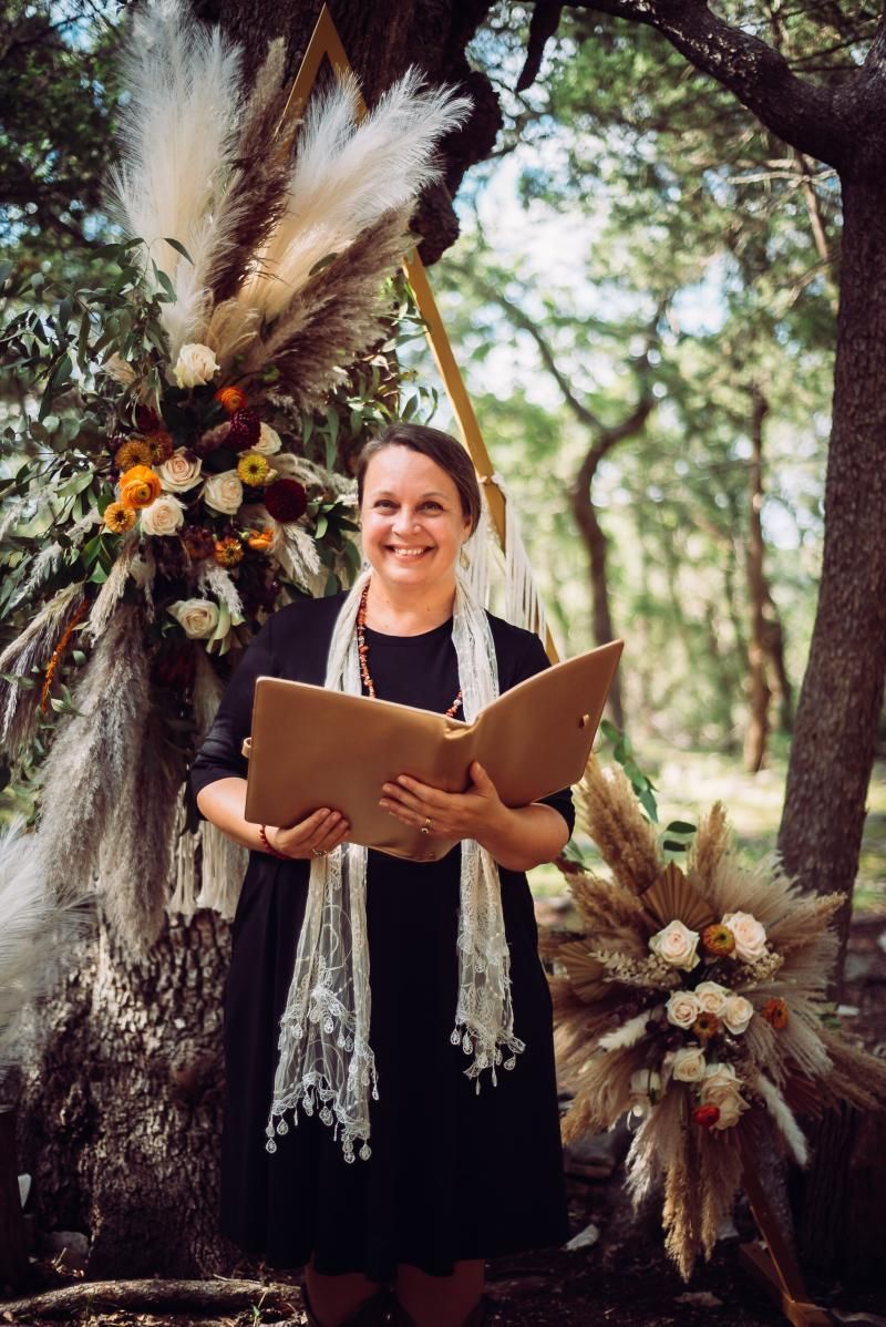 Woman holding an open book in a wooded outdoor setting with a floral arrangement behind her