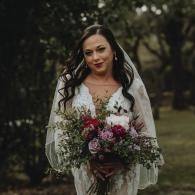 Woman in a white lace dress holding a pink-and-white bouquet outdoors in a wooded setting