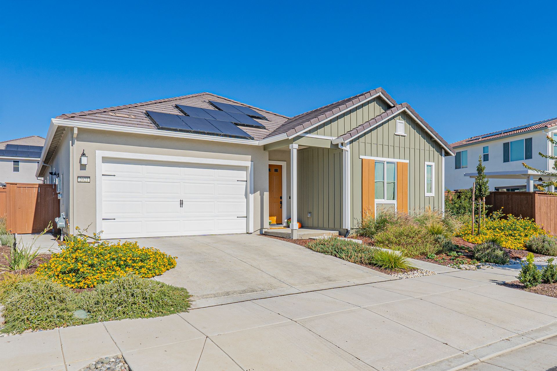 Single-story house with a white garage door, solar panels on the roof, and a green exterior under a clear blue sky.