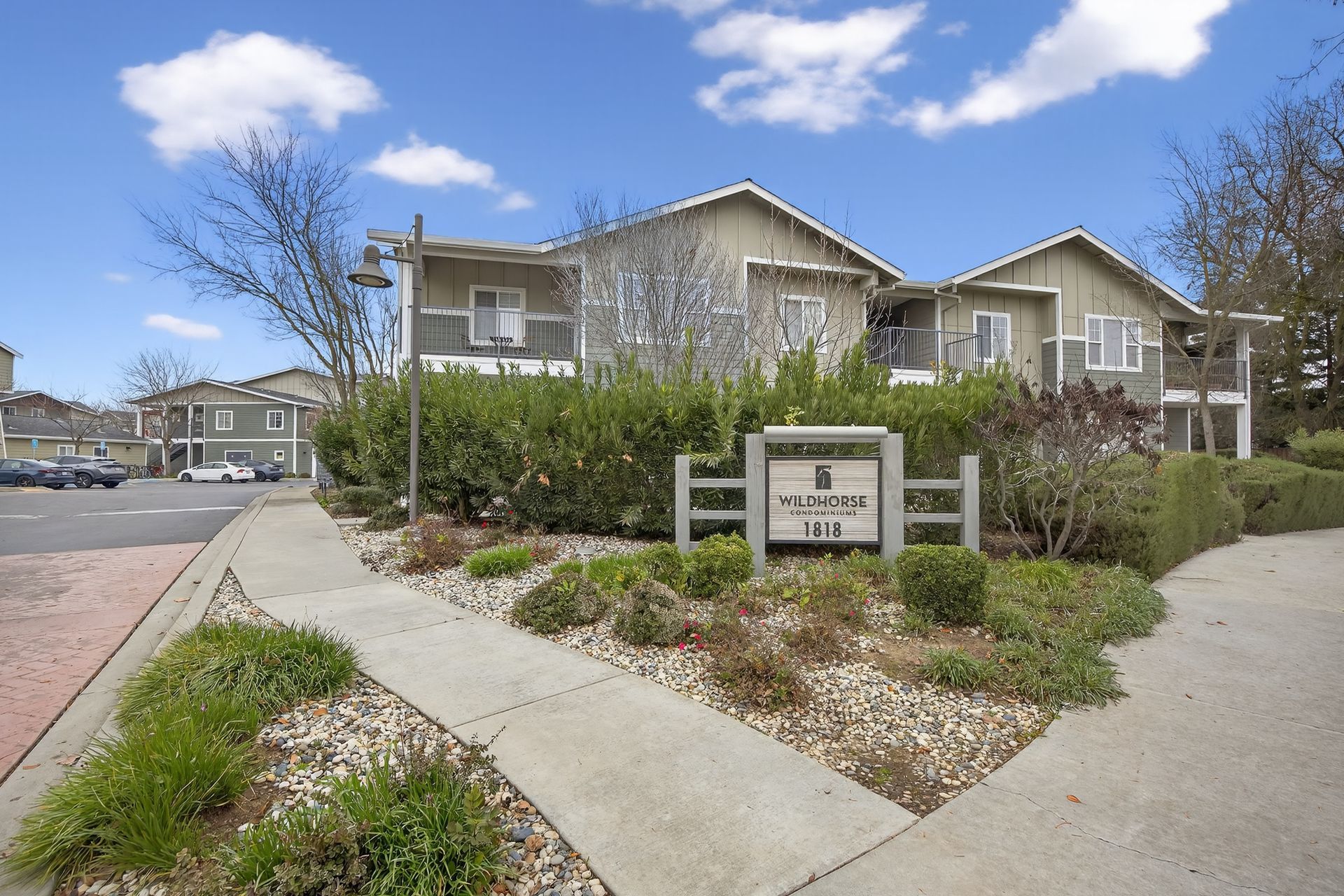 Apartment complex entrance with sign, walkway, and landscaping under a blue sky.