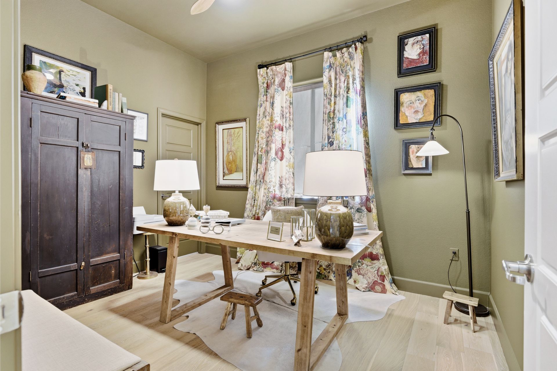 Home office with a wooden desk, floral curtains, artwork, and an armoire.
