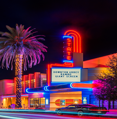 Neon-lit State Theater at night with a palm tree on the left, a car in the foreground, and movie titles on the marquee.