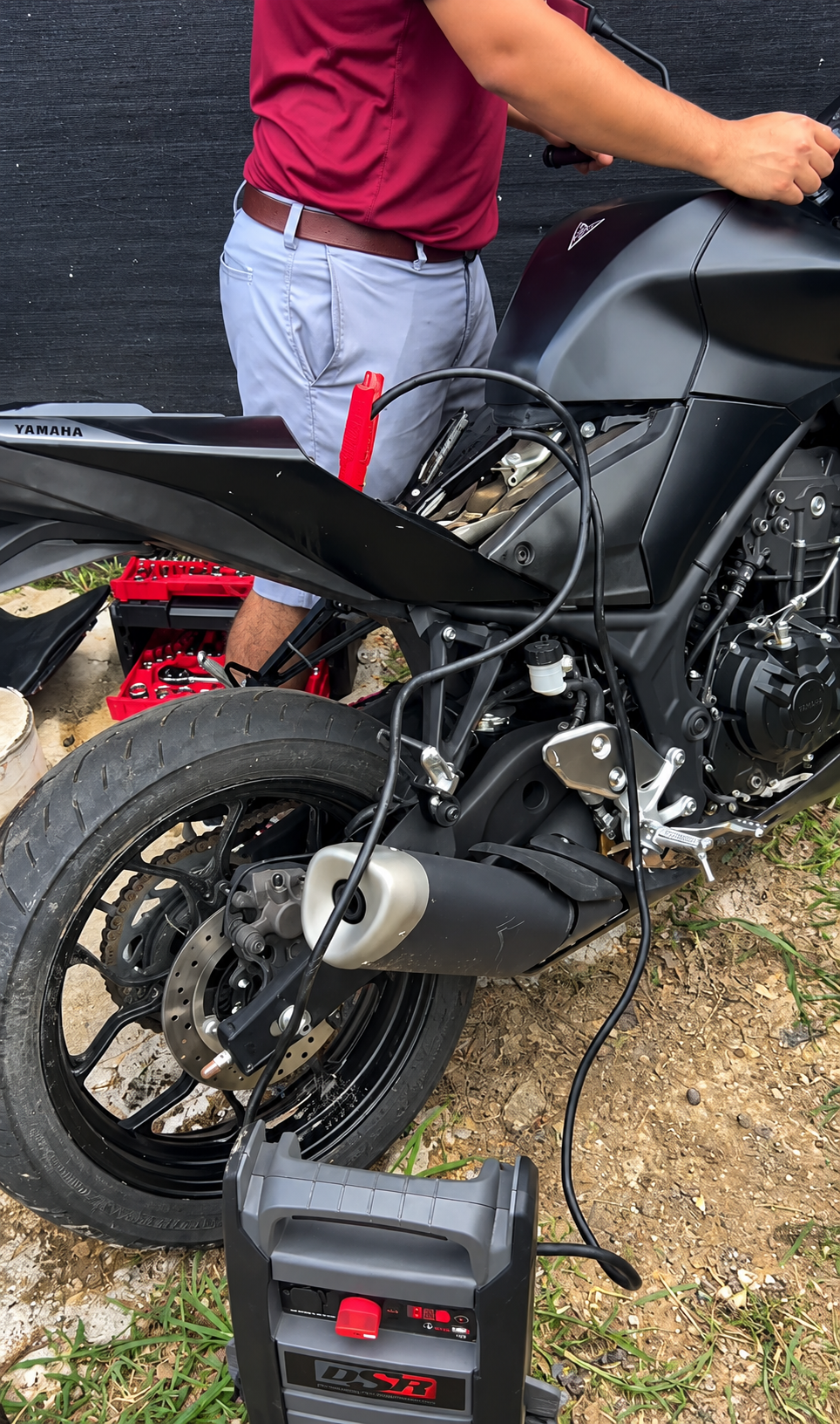 Person standing beside a black motorcycle with a jump starter on the ground nearby