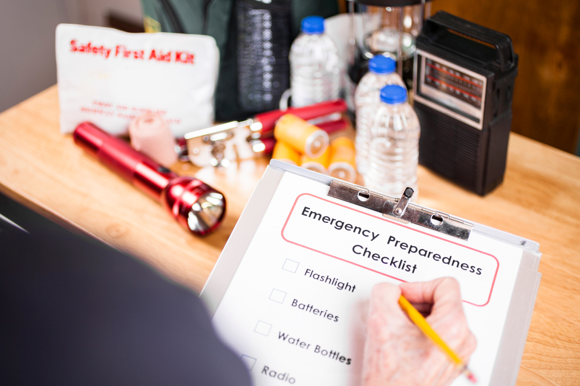 Person checks an emergency preparedness checklist with supplies: flashlight, water bottles, radio.