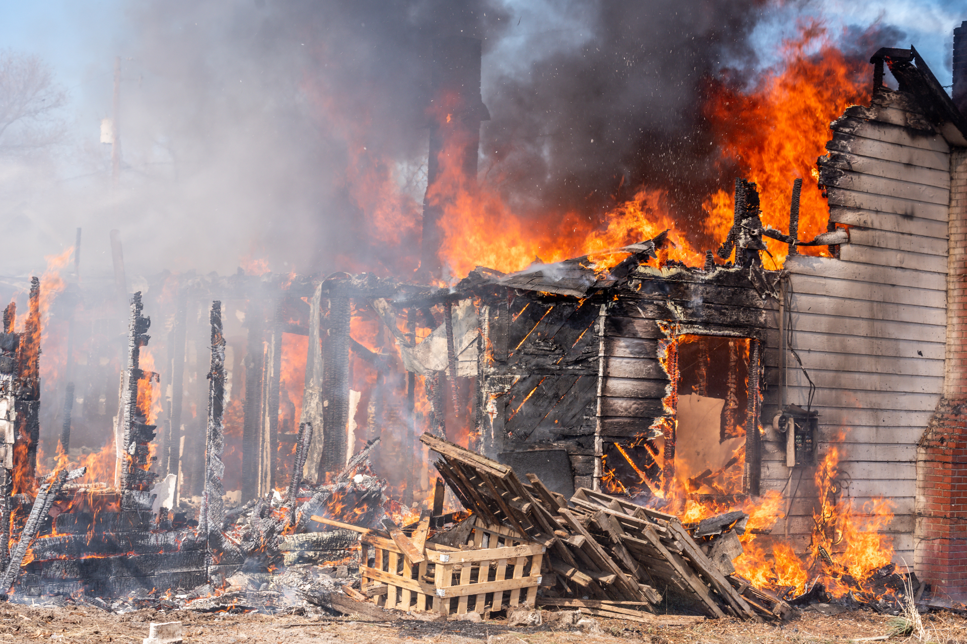 Burning house engulfed in flames, black smoke billowing, charred wood visible.