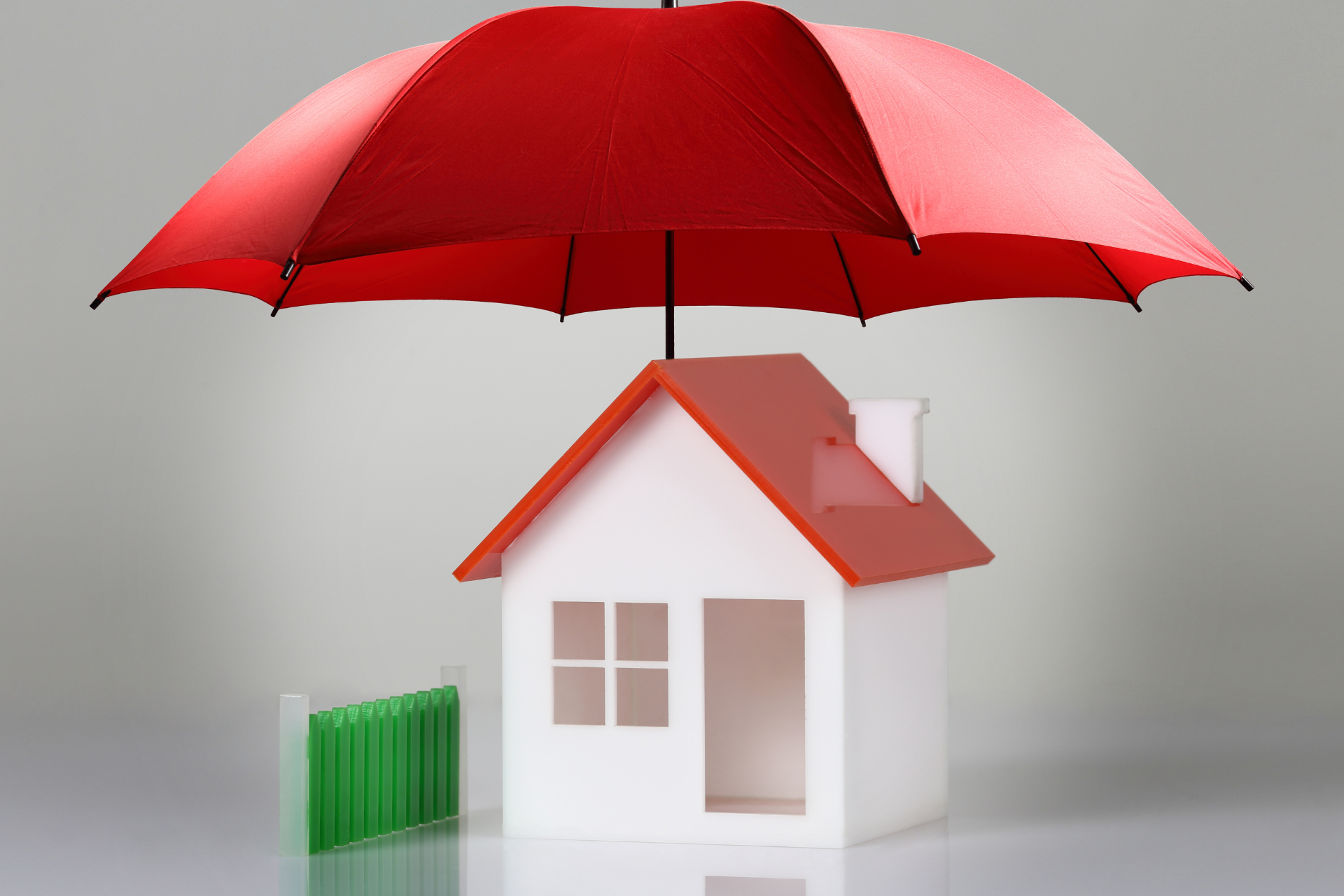 Red umbrella sheltering a small white house with red roof and green fence.