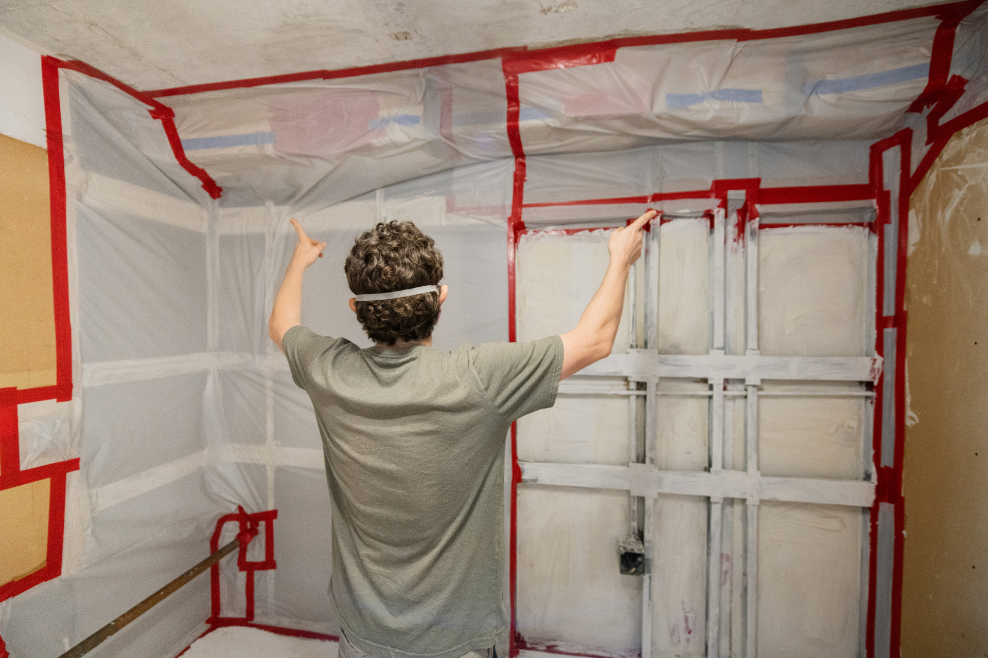 Person inside a room, setting up plastic sheeting with red tape to contain dust.