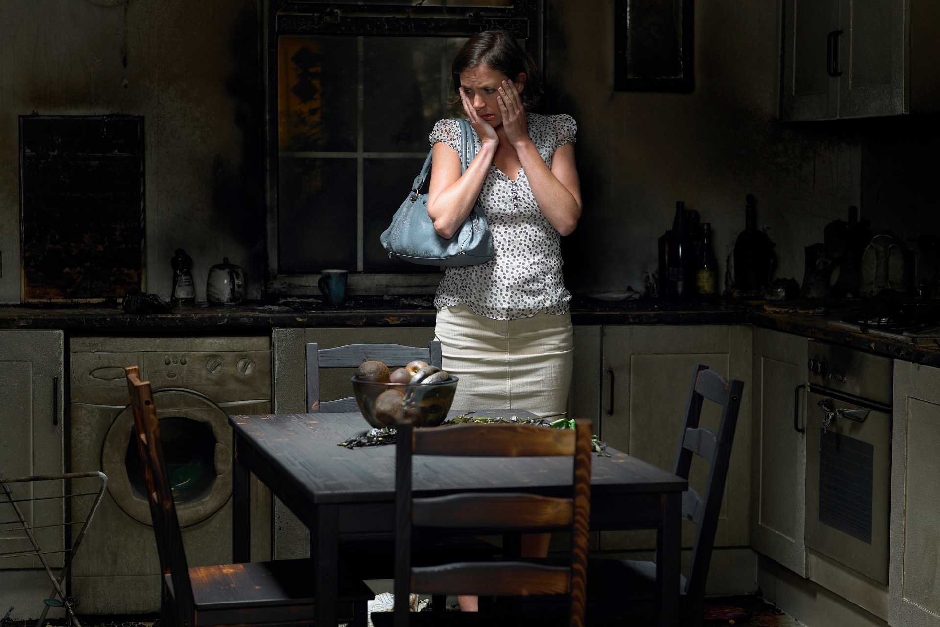 Woman in a burned kitchen, looking distressed. Fire damage visible on cabinets, appliances, and table.