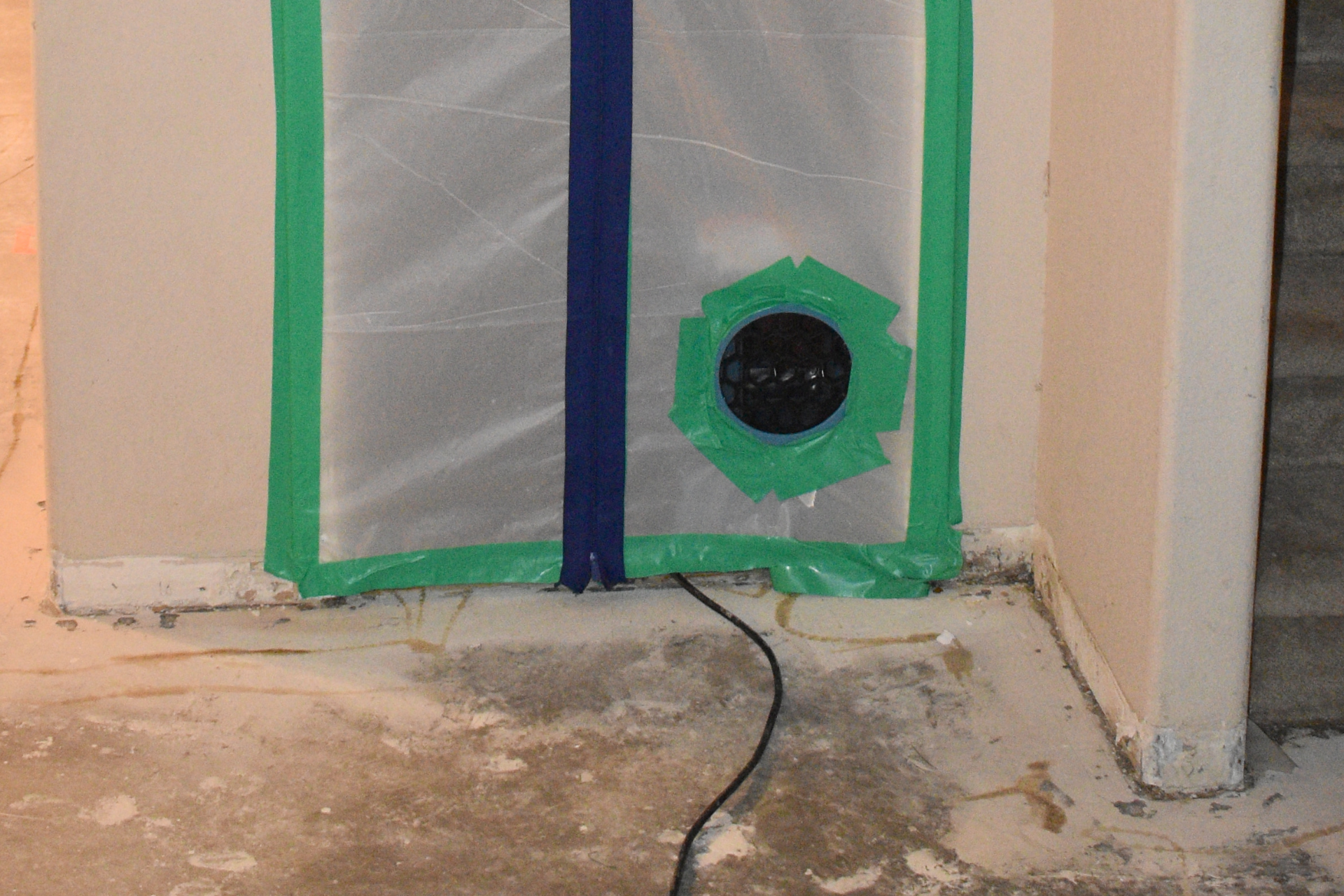 Plastic sheeting sealed with green tape covers a doorway, with a black vent in the center and a power cord below.